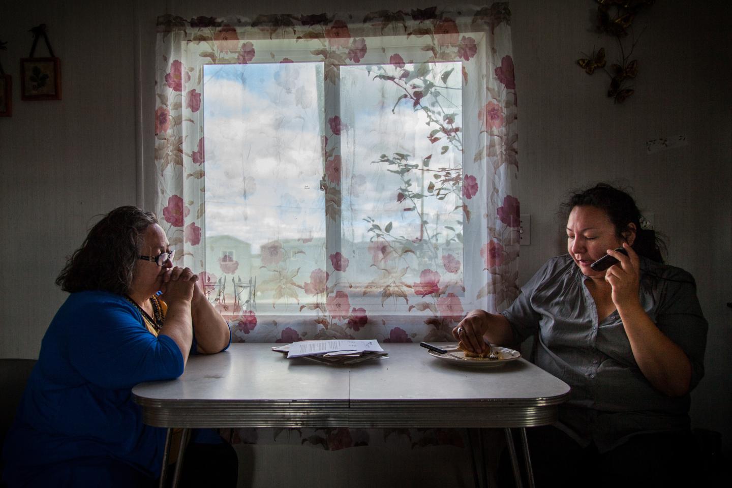 Two Indigenous people sit at a table in front of a window. The person on the right is eating and talking on speaker phone.