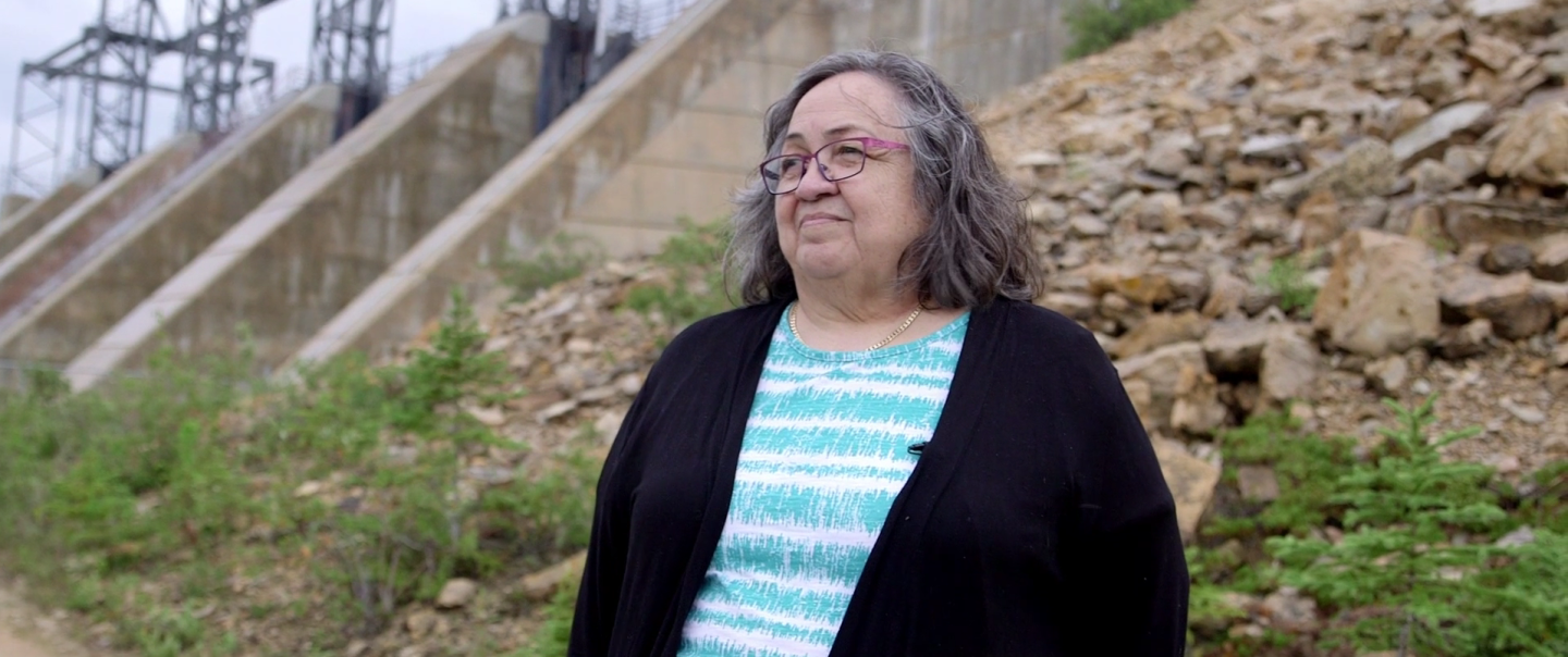 A woman with grey hair and glasses stands in front of a hydro dam