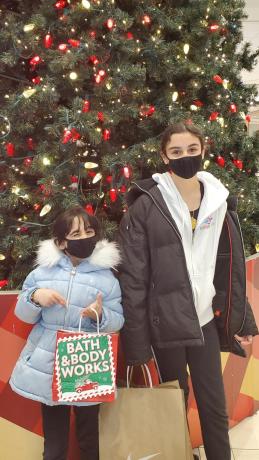 Two girls in winter coats and face masks stand in front of a Christmas tree. They are holding shopping bags.