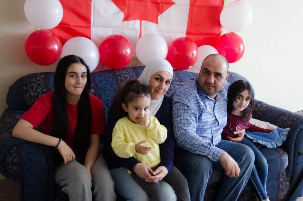 A family of five sits together on a couch. There is a Canadian flag and balloons on the wall behind them.
