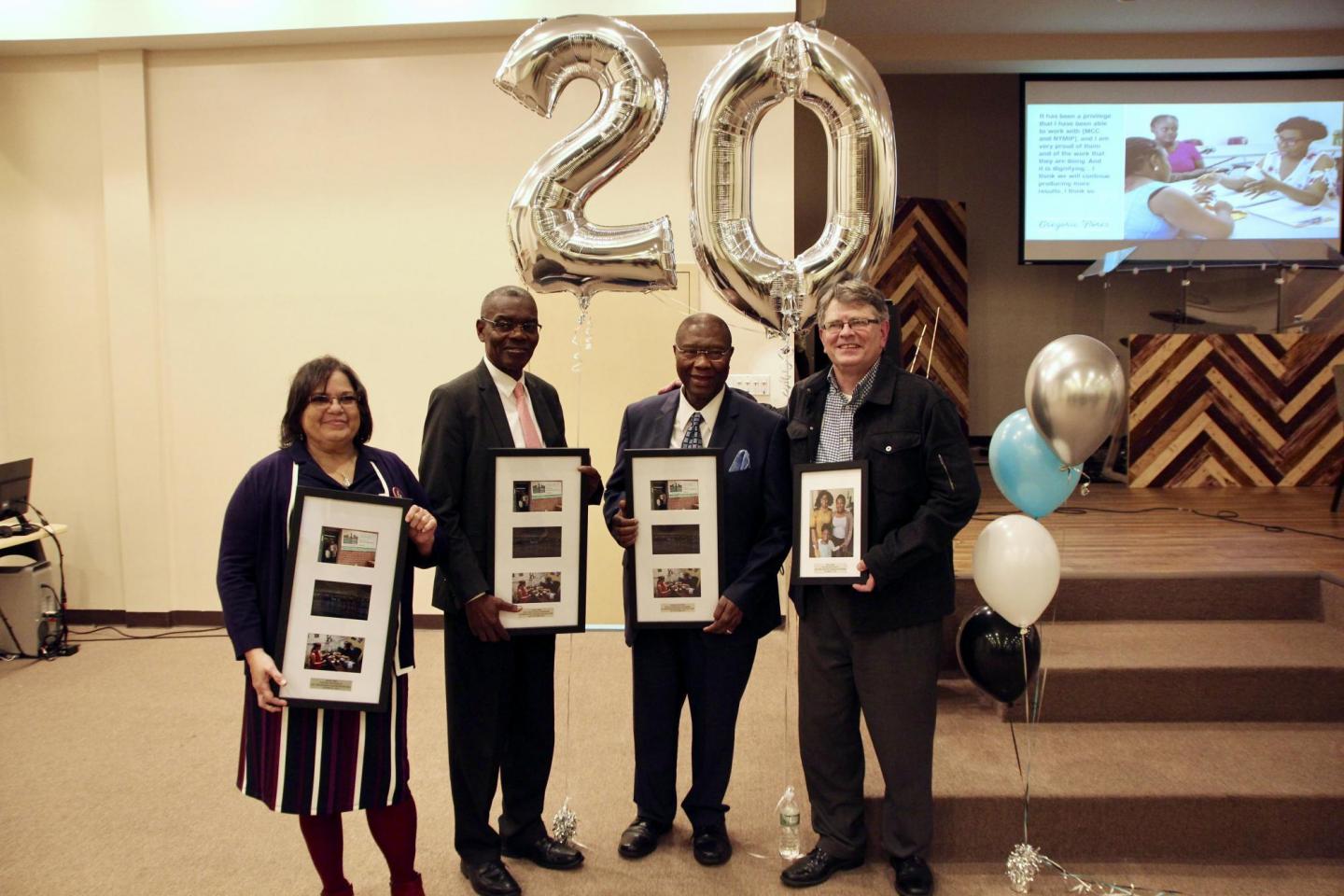 Four people holding picture frames stand together at an event. Behind them are mylar balloons in the shape of the number 20.