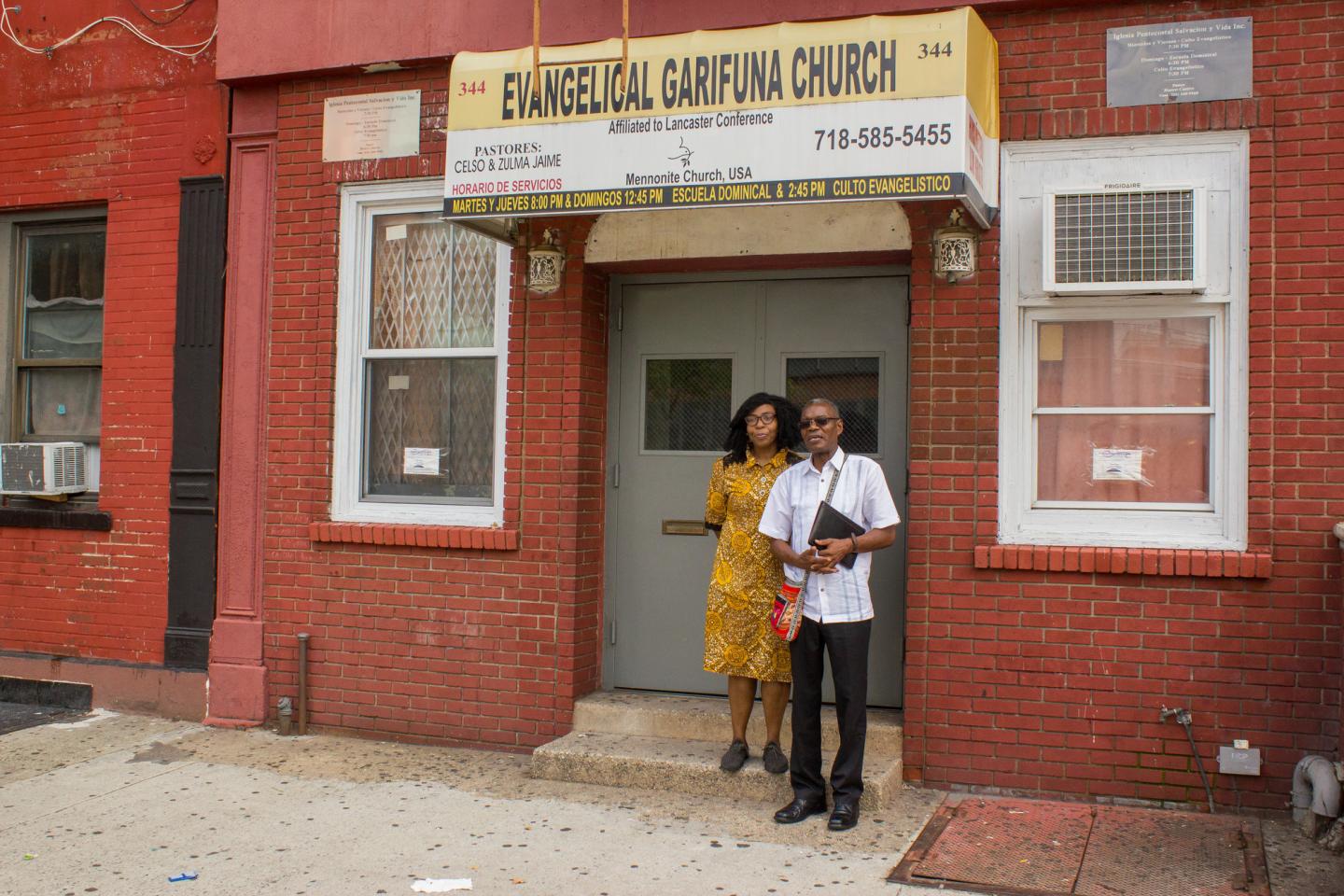 Two people stand by a brick building. The sign on the building reads, "Evangelical Garifuna Church"