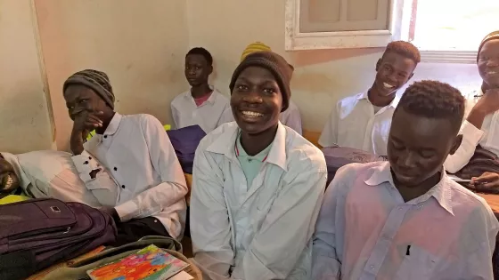 A group of Sudanese students sit at their desks in a small classroom.
