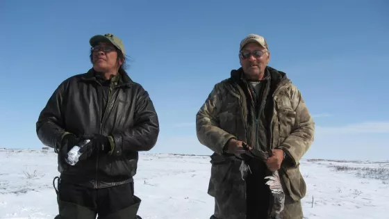 Two men stand with the duck they hunted. There is snow on the ground.