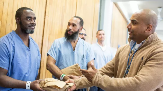 A man in a tan sweater hands a paper bag to another man in blue prison clothes