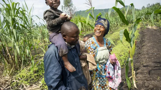 A family of four in the DR Congo walk together in a field. A child is on the father's shoulders laughing and eating