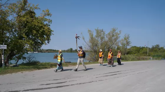 A group of people walk with a pail of water and an eagle staff alongside a river