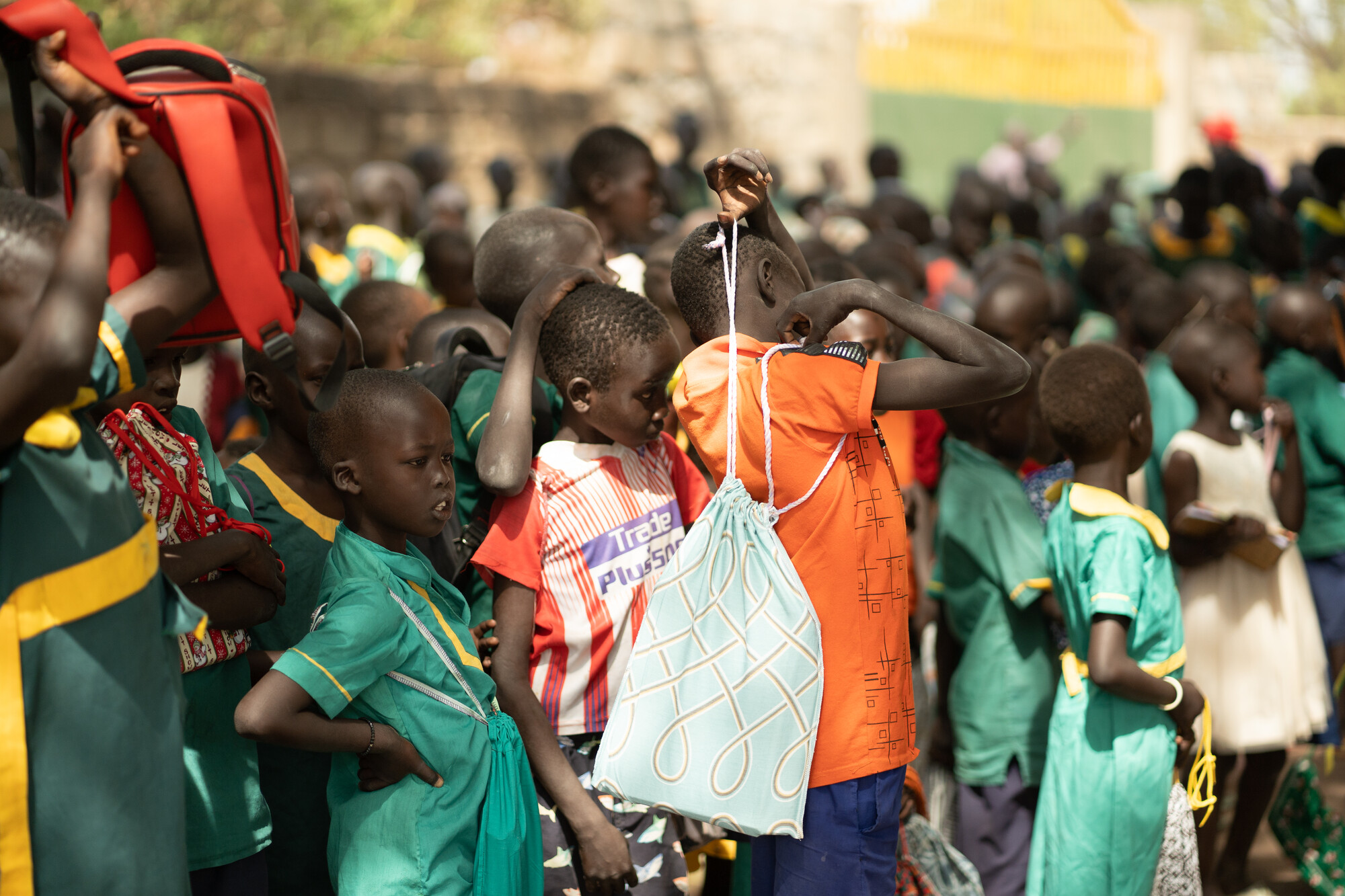 A large group of South Sudanese students. A child in the center in an orange shirt has a cloth bag thrown over his shoulders
