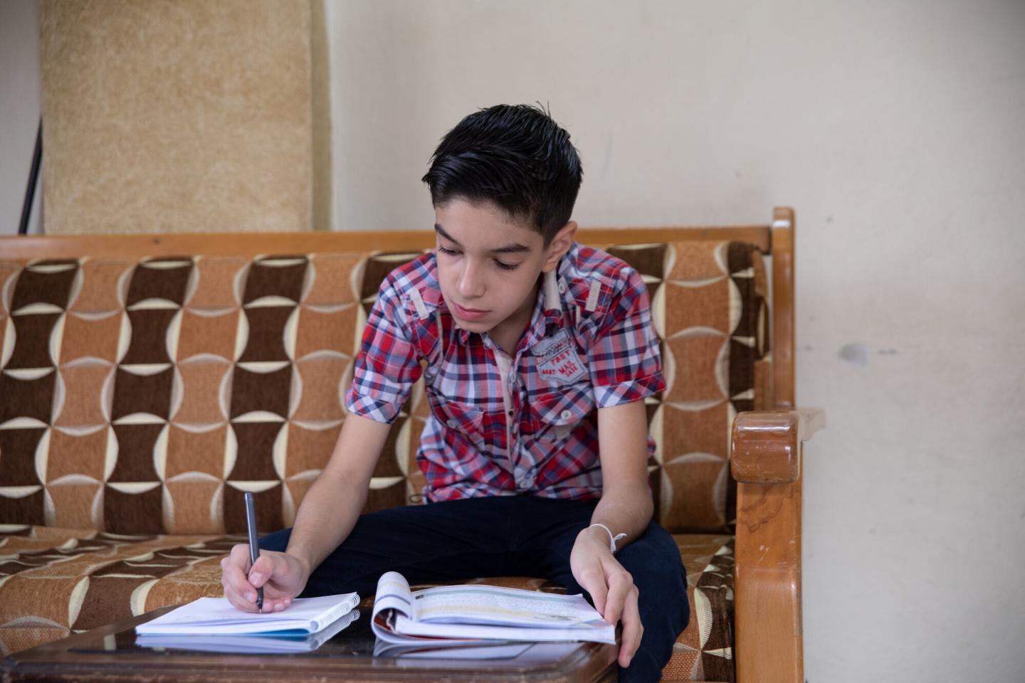 A Syrian boy sits on a couch and leans over his school work