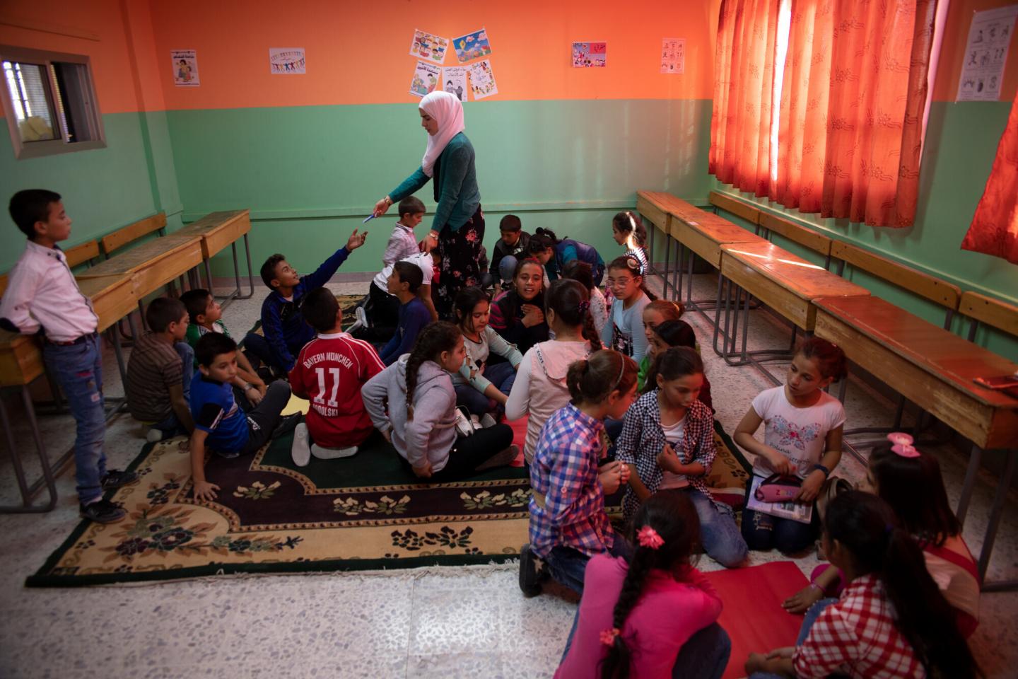A Syrian teacher hands something to a child sitting among a group of children on a rug.