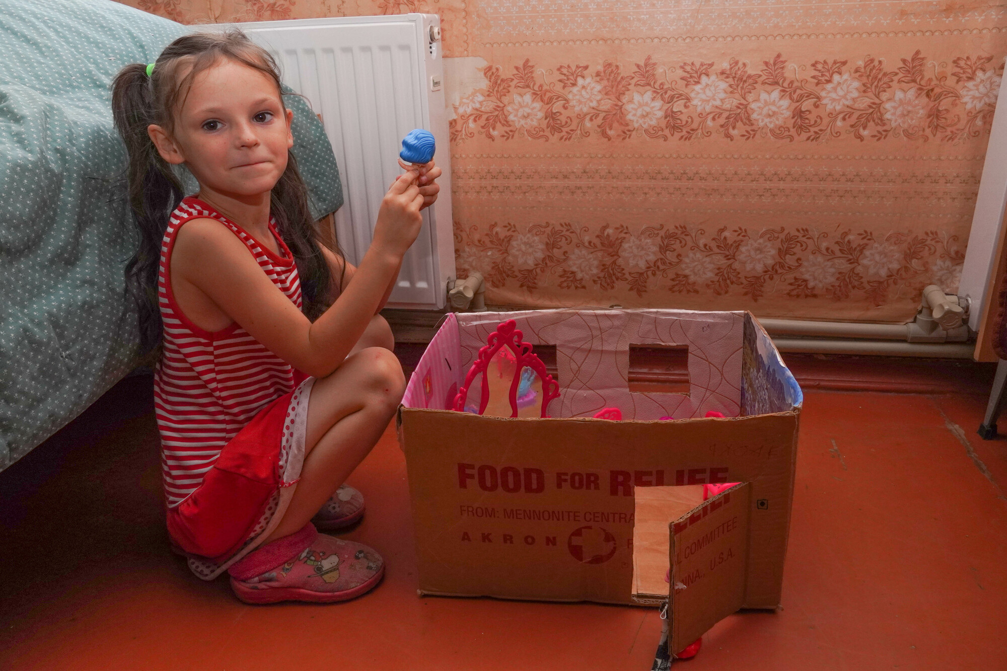 A young girl playing with a doll beside a dollhouse made of an MCC canned meat box
