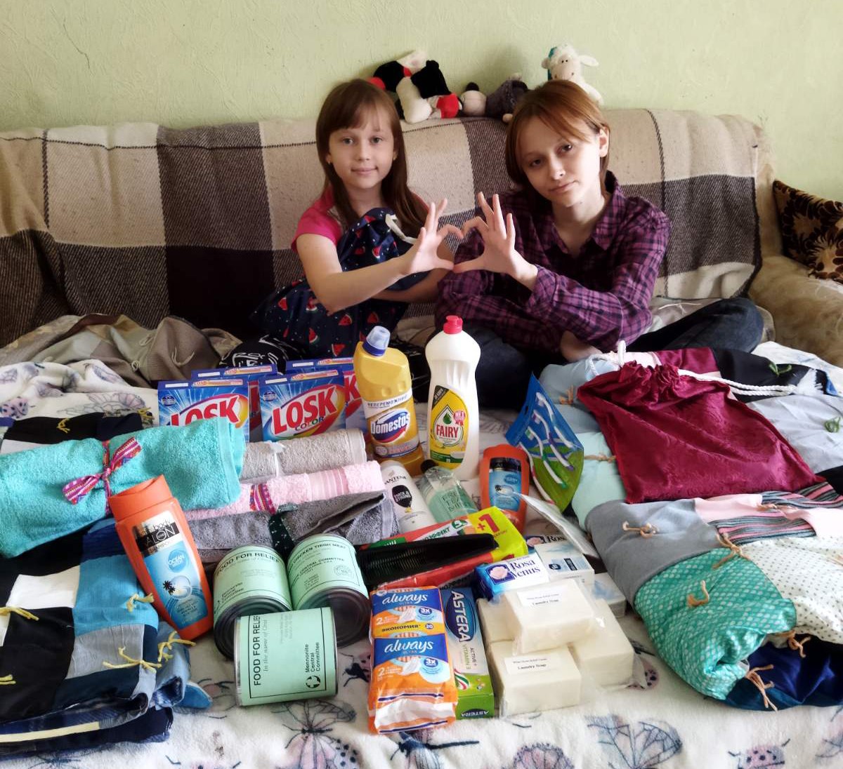 Two girls making a heart symbol with their hands sitting in front of relief supplies on a bed