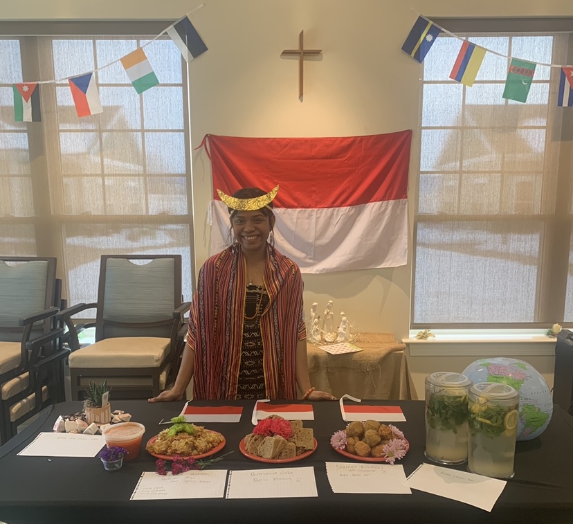 A young woman in traditional Indonesian clothing stands behind a table with traditional food