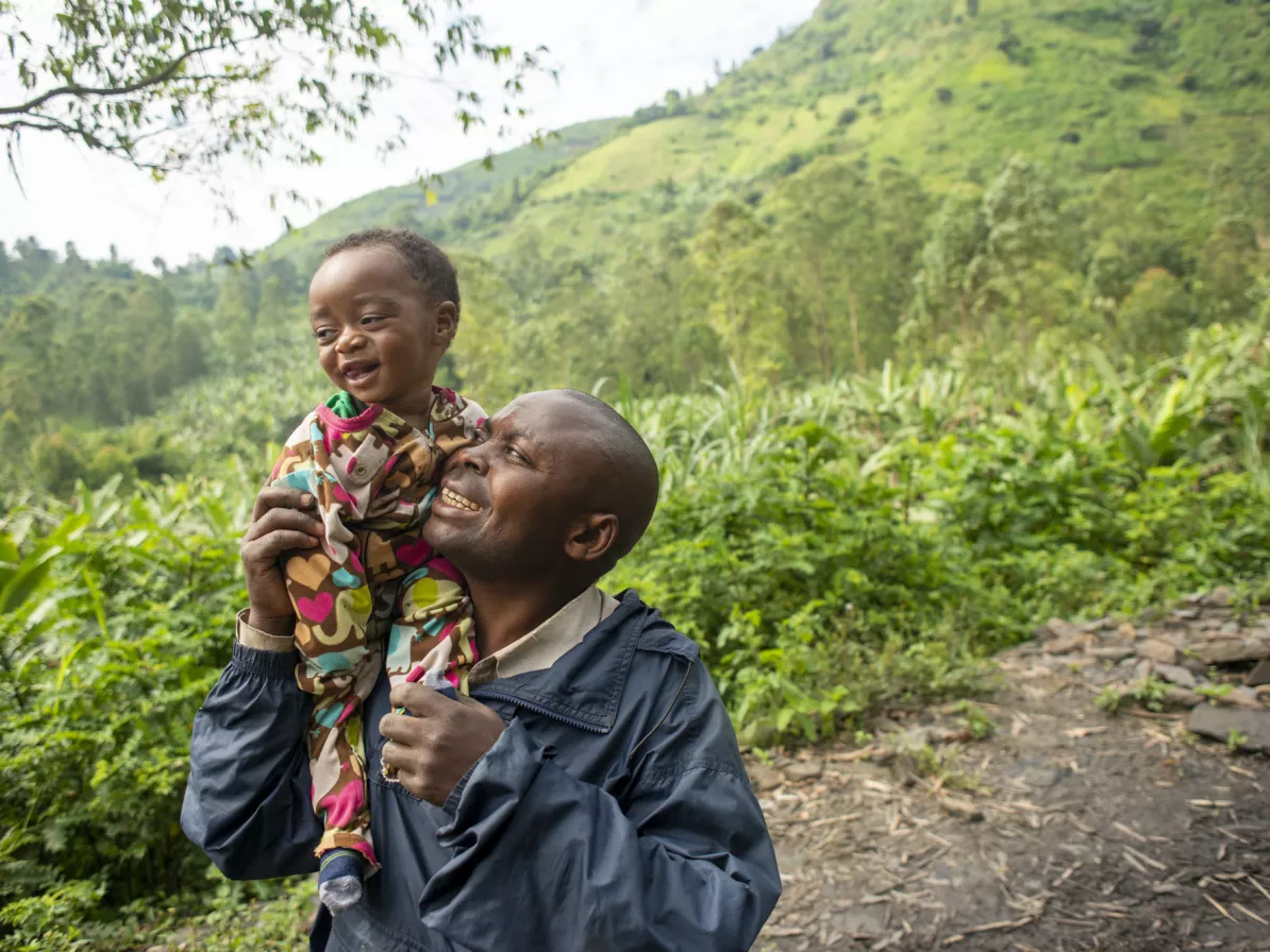 A father holds his child on his shoulder and smiles up at them. They are near a field in a DR Congo.